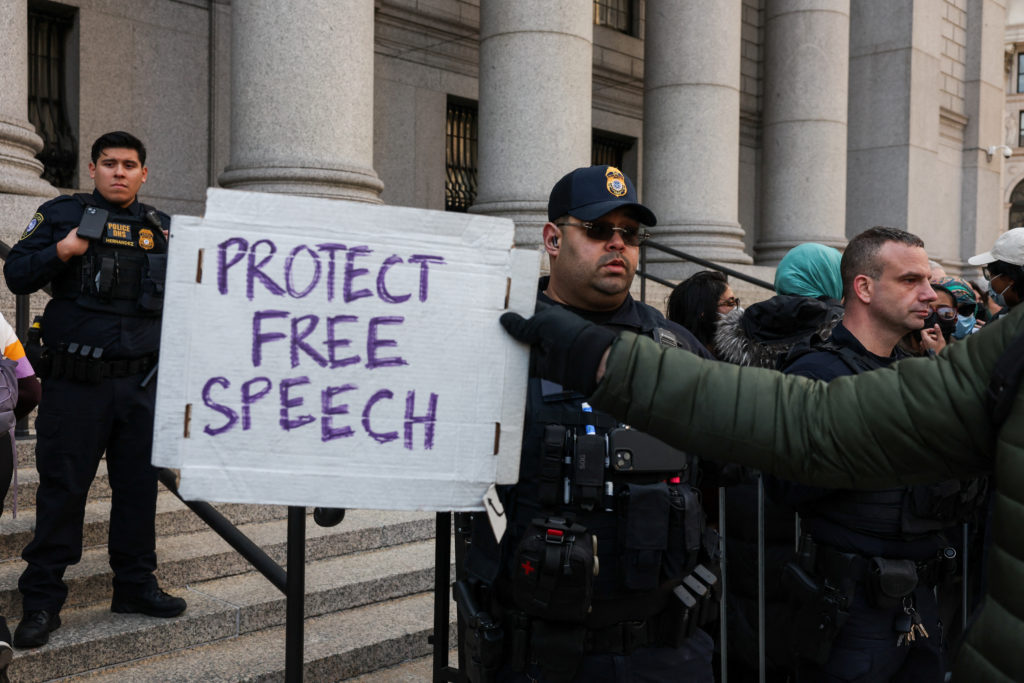 Protester holding “I Protect Free Speech” sign at a Trump-related event with security present.
