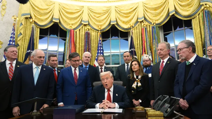 A group of officials and advisors stand around a seated U.S. president in the Oval Office as he signs a document.